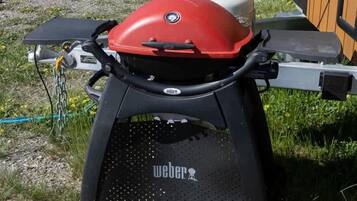 Una parrilla portátil con parte superior roja, base negra y ruedas, ubicada sobre césped cerca de una estructura de madera.