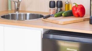 Una cocina con un lavamanos de acero inoxidable, una encimera de madera, una ventana con vistas a árboles, un lavavajillas Hisense y una tabla de cortar con verduras y molinillos de pimienta.
