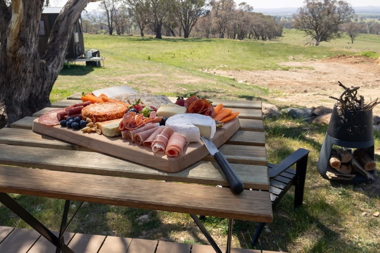 Un pique-nique avec une variété de fromages, de charcuteries et de fruits, disposés sur une table en bois dans un cadre extérieur.