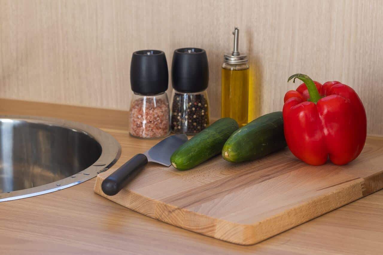 A kitchen counter with a cutting board, a knife, a red bell pepper, cucumbers, and two pepper mills.