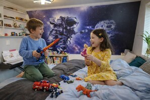A child and an adult playing with toys on a bed with a space-themed wall mural.