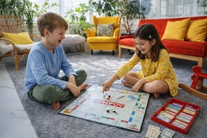 Two children playing a board game on a carpeted floor in a room with a red sofa and yellow chairs.