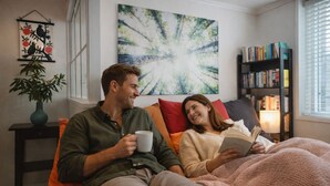 Un couple est assis sur un lit, avec une bibliothèque et un tableau en arrière-plan.