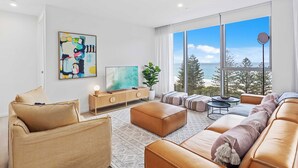 A modern living room with a large window, a flat-screen TV, and a sofa set. - Luxury Ocean View Burleigh Apartment (Burleigh Heads)