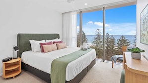 A modern bedroom with a large bed, a bedside table with a lamp, a small wooden side table, a desk with a plant, and a view of the ocean through large windows. - Luxury Ocean View Burleigh Apartment (Burleigh Heads)