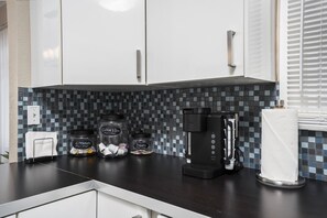 Cocina con gabinetes blancos, cafetera y tarros etiquetados como 'Café Té', 'Edulcorantes' y 'Azúcares'.