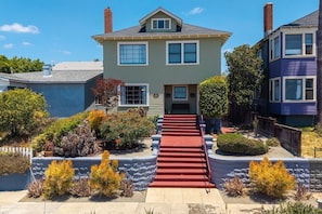 A two-story house with a red carpet leading to the front door, surrounded by landscaped gardens and a white picket fence.