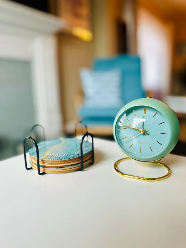 A teal-colored alarm clock with a gold frame on a white surface, next to a stack of coasters with a blue and white design.