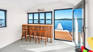 A modern interior with a wooden bar and stools, large windows offering a view of the sea, and a sign that reads "Simply Blessed". - Catalina Bay - Absolute Lakefront - Jetty - Boat house on waters edge (Fishing Point)