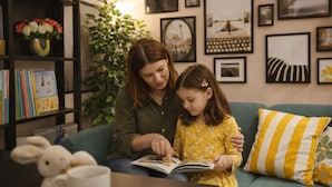 Una mujer y un niño leyendo un libro juntos en un sofá, con una estantería y almohadones decorativos al fondo.