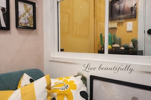 A room with a window, a bed with a yellow and white pillow, and a framed picture with the text "Live beautifully."