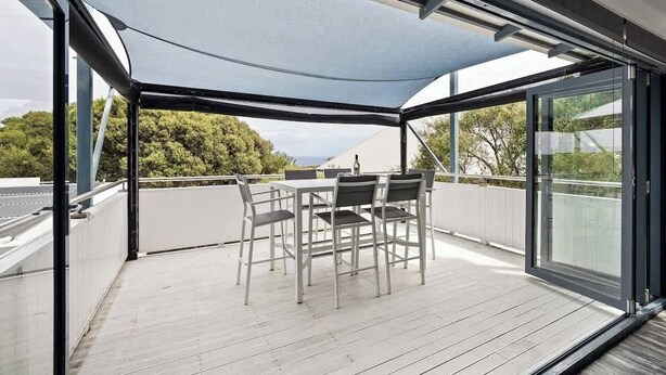 A covered outdoor seating area with a table and chairs, large windows, and a view of greenery. - Surfers Paradise—Oceanview Garden Beach House (Yallingup)