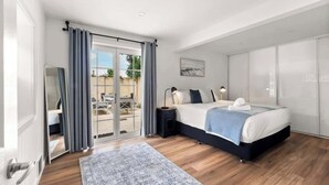 A bedroom with a bed, a nightstand, a lamp, a window with curtains, and a sliding door leading to an outdoor area. - Surfers Paradise—Oceanview Garden Beach House (Yallingup)