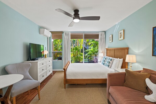 A hotel room with a bed, a TV, a desk, a chair, a sofa, and a balcony with a view of greenery.