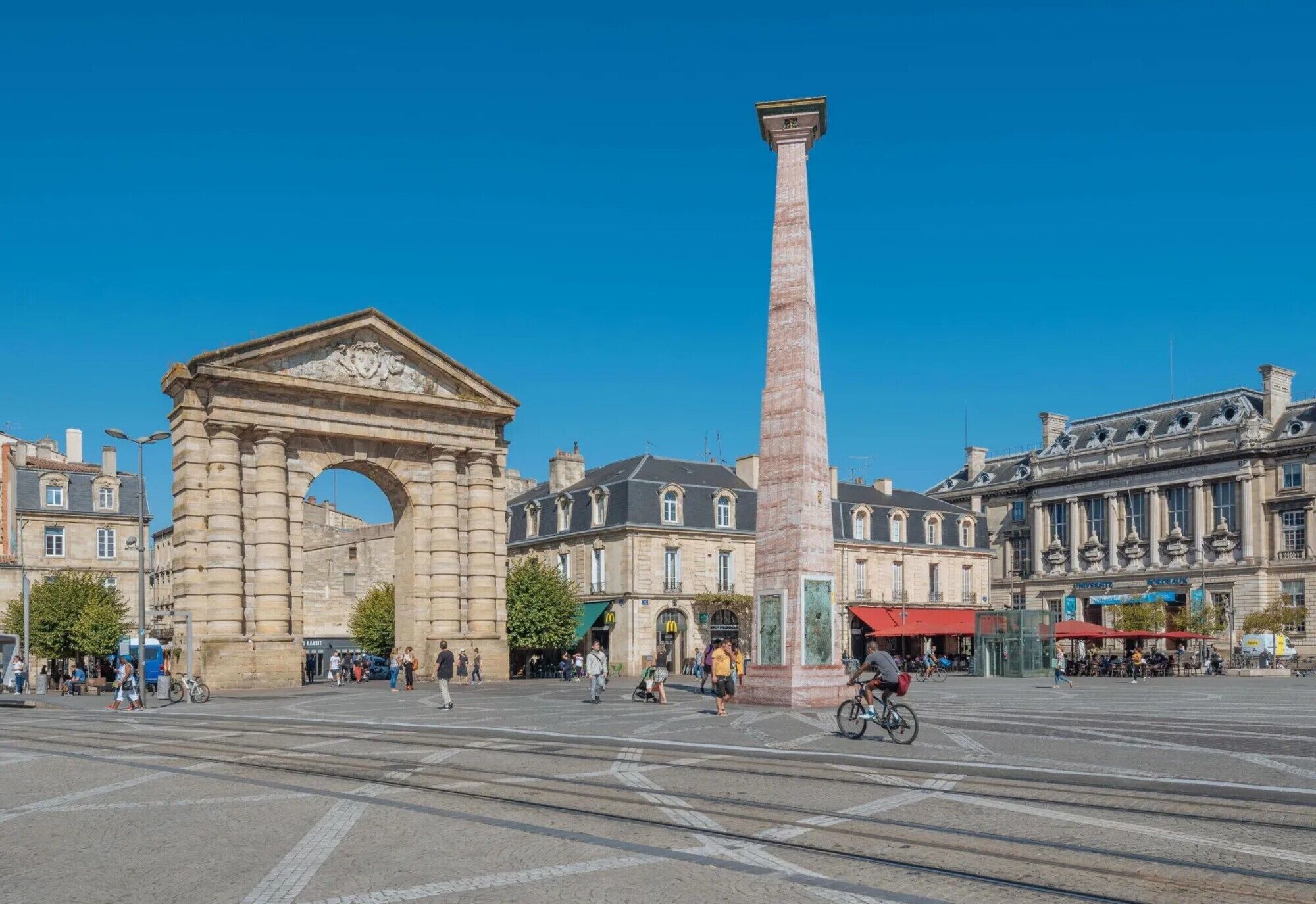A historic square with a prominent archway and a tall column, surrounded by classical buildings.