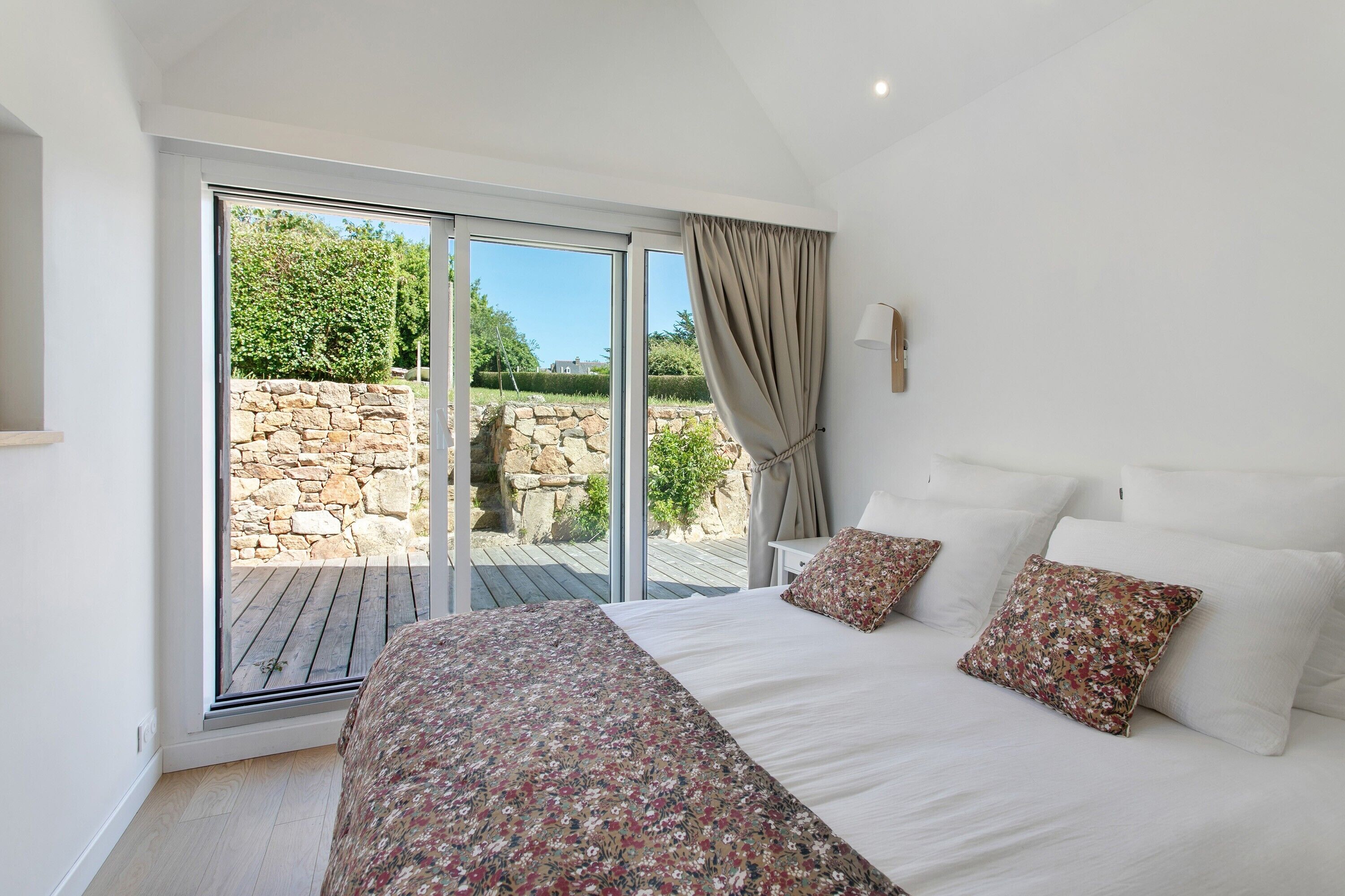 A bedroom with a large sliding glass door leading to a patio, a bed with floral bedding, and a stone wall in the background.