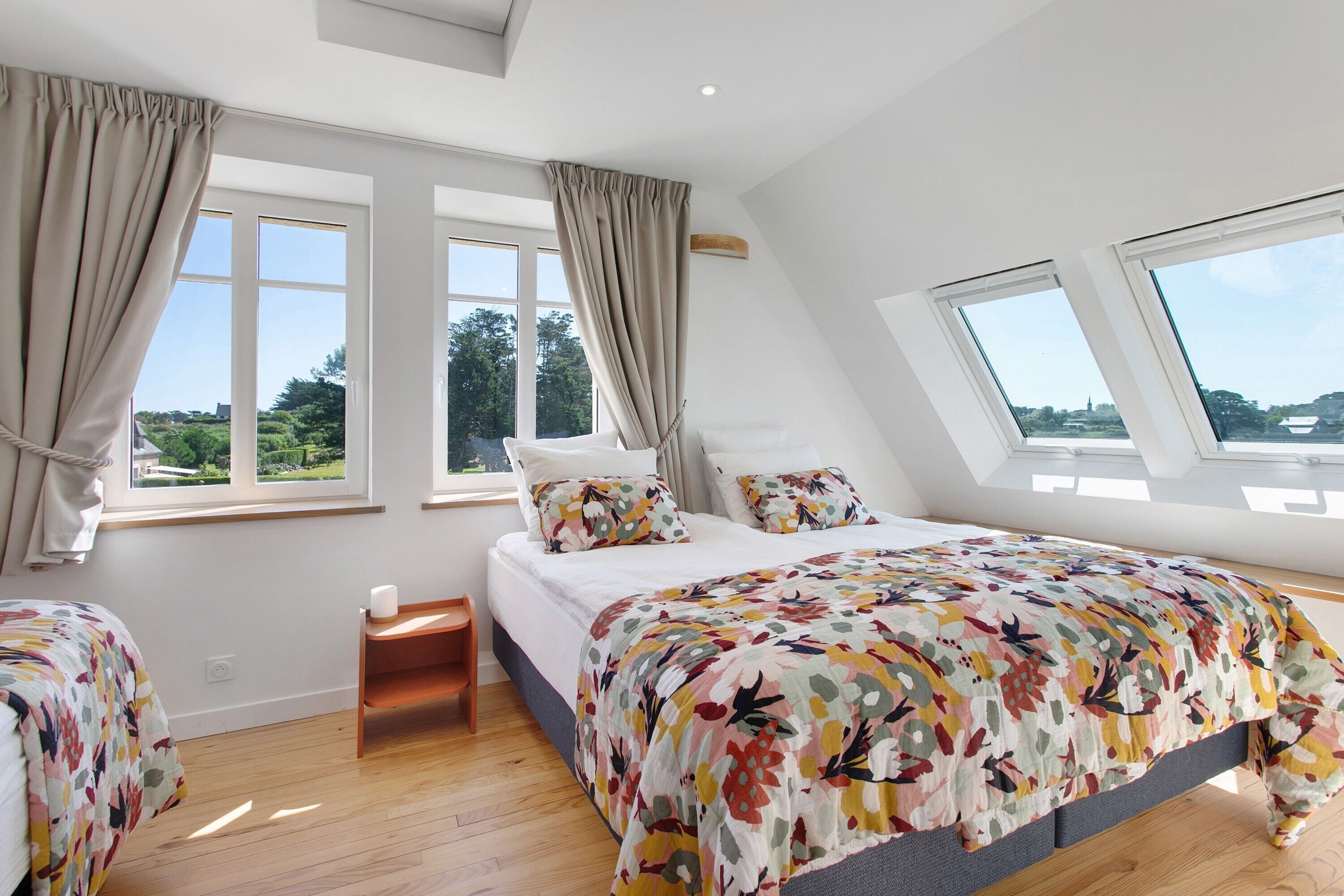 Un dormitorio con cama, dos ventanas, tragaluz y vistas al exterior con vegetación.