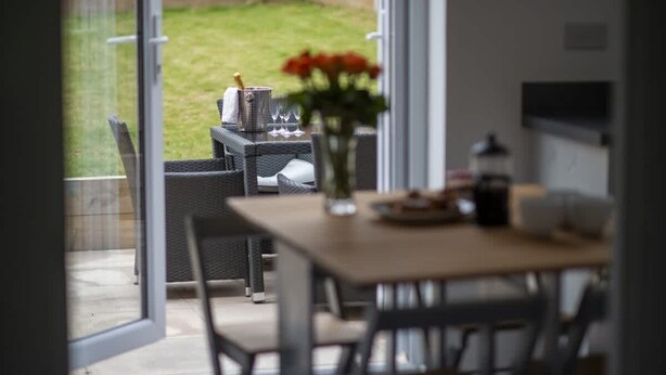 Un'area pranzo con tavolo e sedie, un vaso con fiori, e vista su un giardino retrostante con barbecue e mobili da esterno.