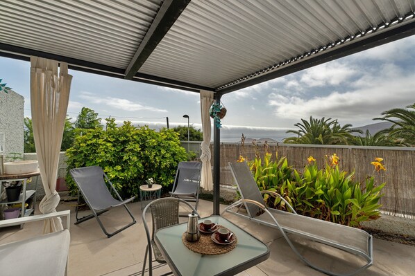 A covered patio with two lounge chairs, a table with a teapot and cups, and a view of greenery and mountains.