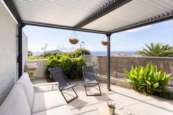 En overdækket terrasse med hvidt loft, to sorte liggestole, en hvid sofa og en potteplante.