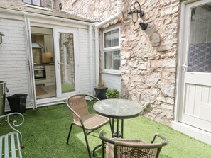Un petit espace patio avec une table ronde en verre et des chaises en osier, un mur en pierre et une porte blanche donnant sur une cuisine.