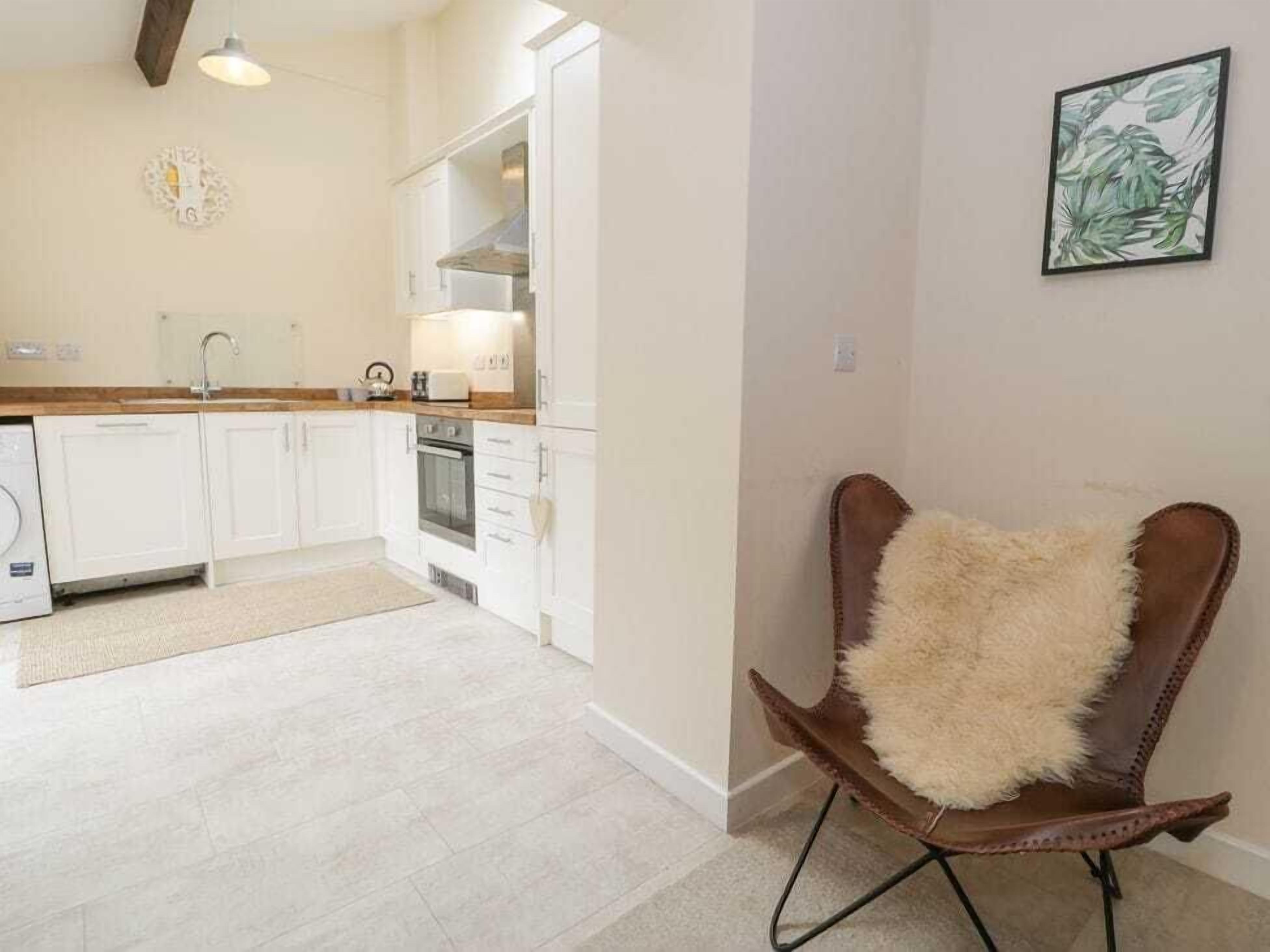 A modern kitchen with white cabinets, a wooden countertop, and a built-in oven. There is a fluffy chair with a brown leather seat and a white cushion.