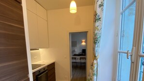 A narrow hallway with a kitchen on one side, a dining area in the background, and a window with curtains on the other side.