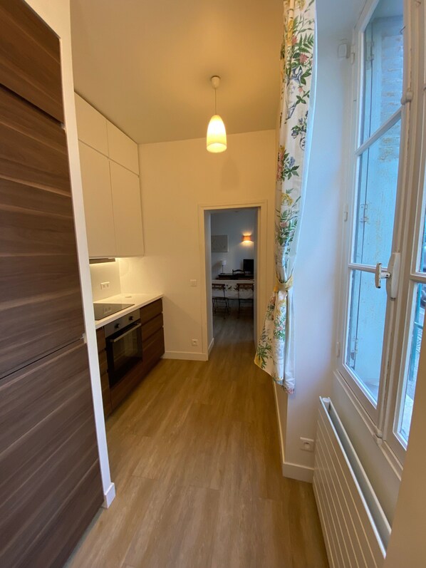 A narrow hallway with a kitchen on one side, a dining area in the background, and a window with curtains on the other side.