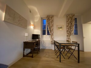 A room with a wooden desk, a television, a dining table with chairs, a staircase, and a door.