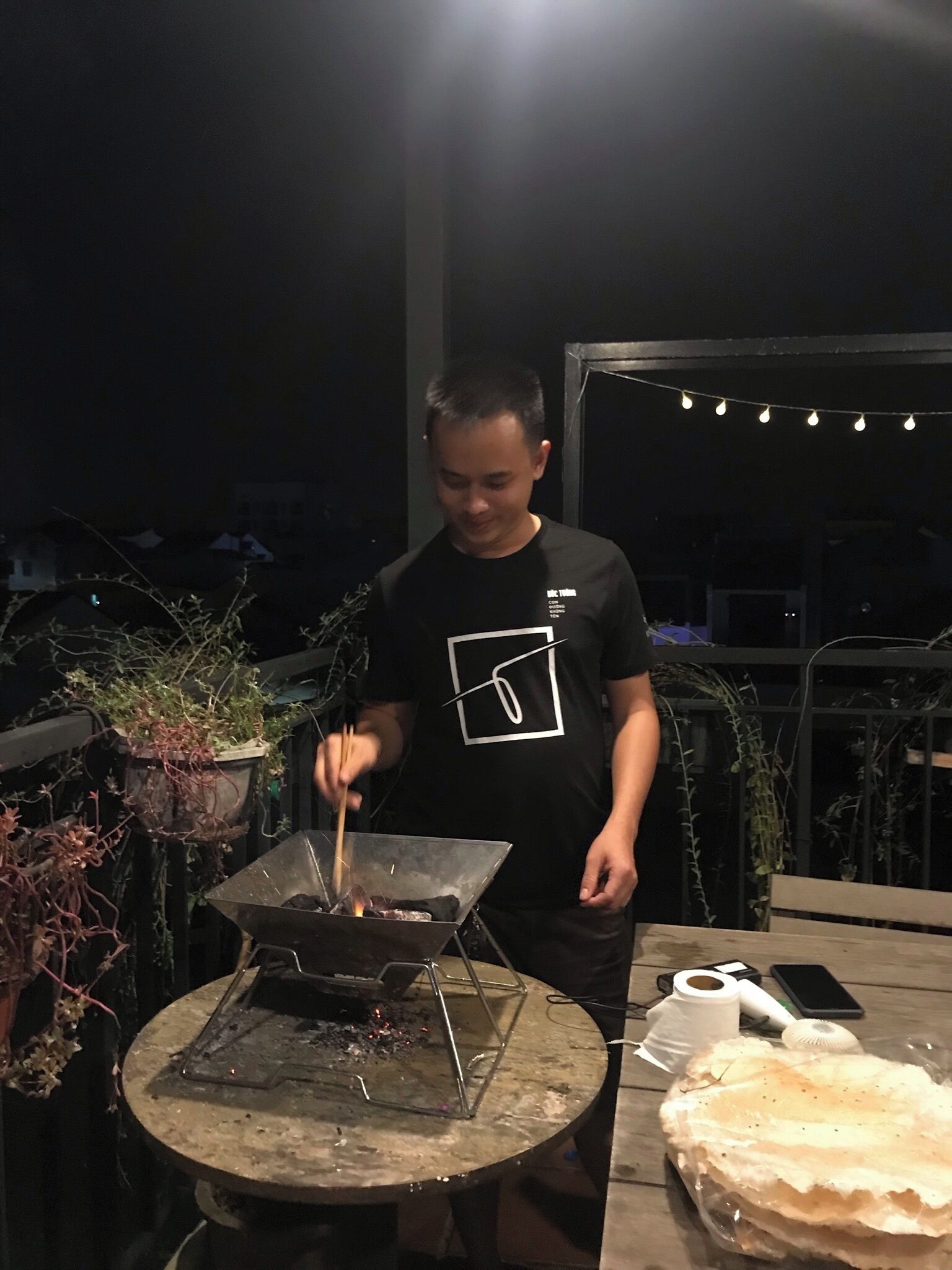 Une personne cuisine à l’extérieur, la nuit, à l’aide d’un réchaud portable, entourée de plantes en pot et de guirlandes lumineuses.
