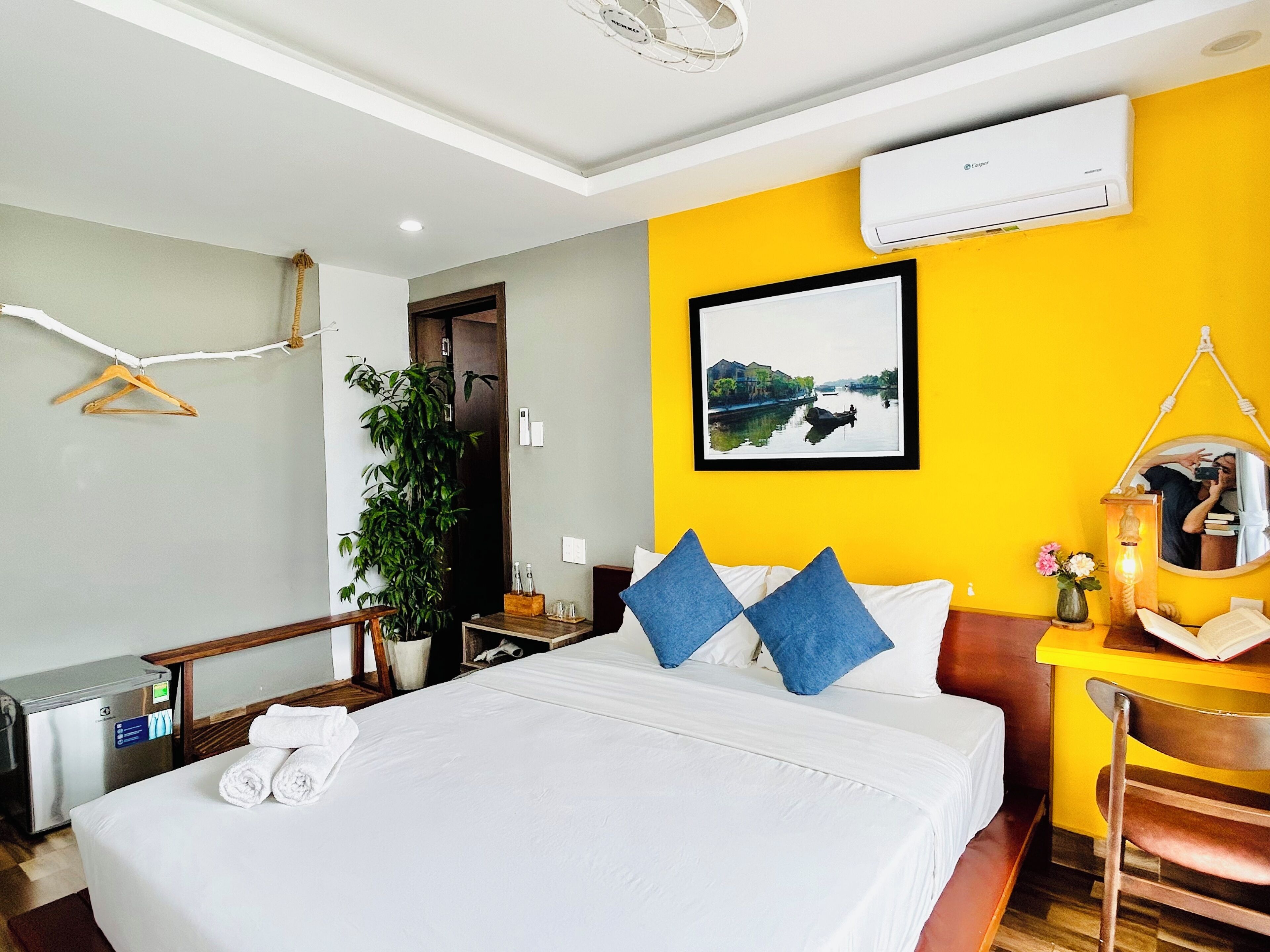 A hotel room with a bed, a desk, a chair, a plant, and an air conditioning unit.