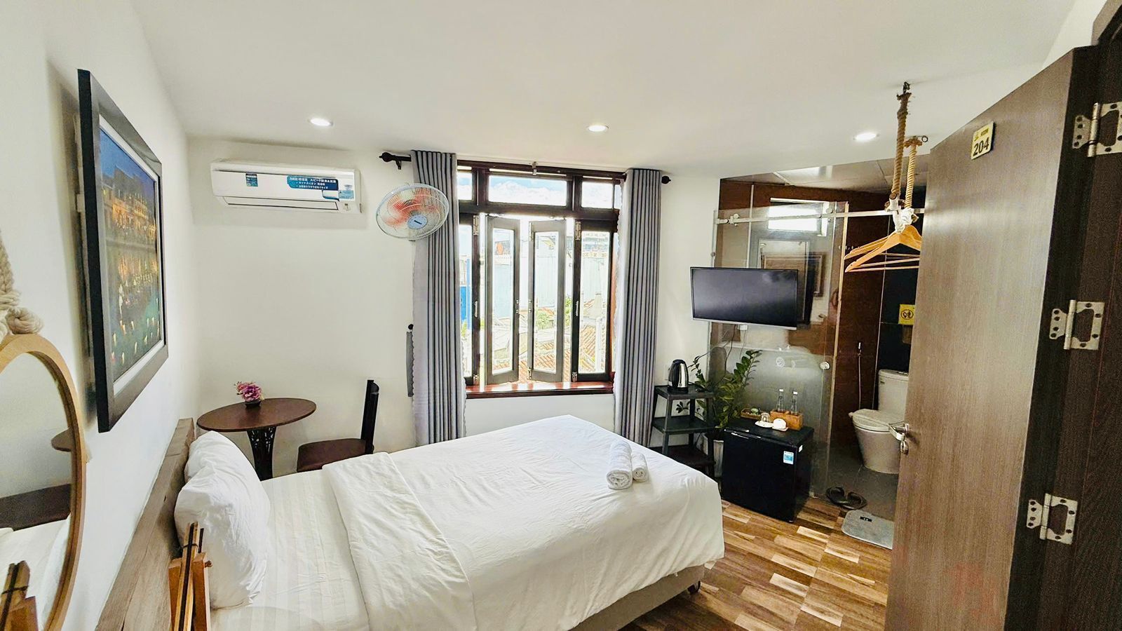 A hotel room with a bed, a TV, a fan, a small table, and a window with curtains.