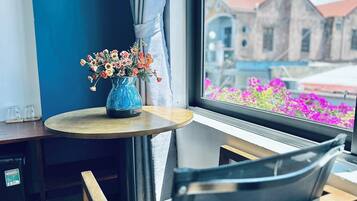Habitació amb vistes a edificis i flors, un ram de flors en un vas blau sobre una taula petita i una cadira.