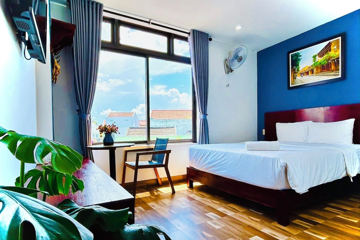 A hotel room with a bed, a desk with a chair, a window with curtains, a plant, and a painting on the wall.