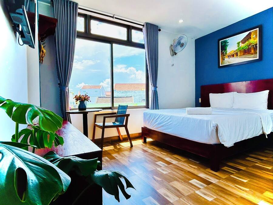 A hotel room with a bed, a desk with a chair, a window with curtains, a plant, and a painting on the wall.