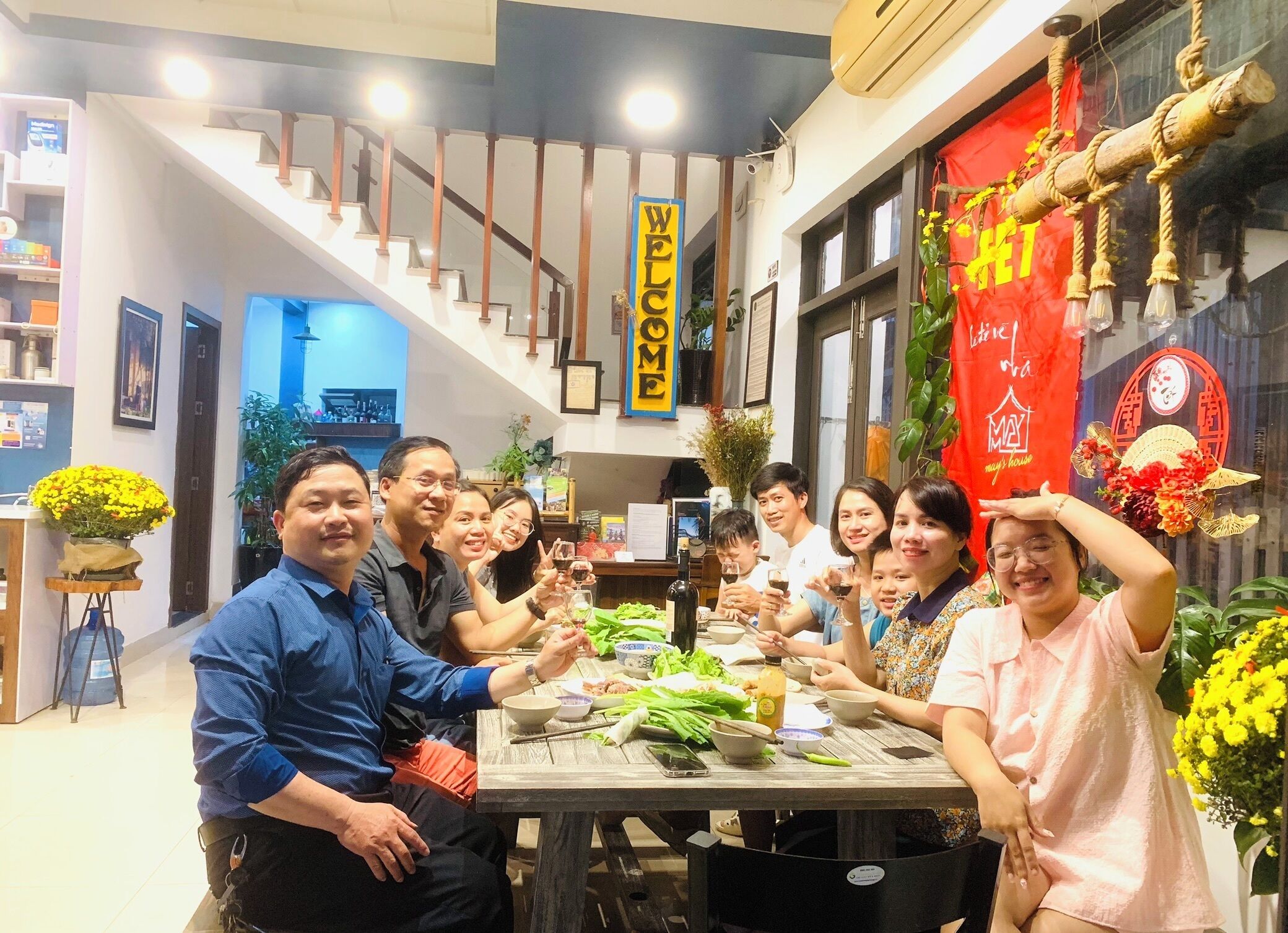 Un grupo de personas sentadas alrededor de una mesa con comida y bebidas, en una habitación con una escalera y un cartel que dice 'WELCOME'.