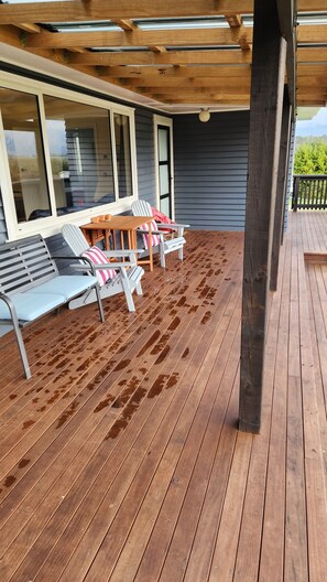 Eine Holzterrasse mit überdachter Sitzecke, inklusive Tisch und Stühlen, und Blick auf das Haus im Hintergrund.