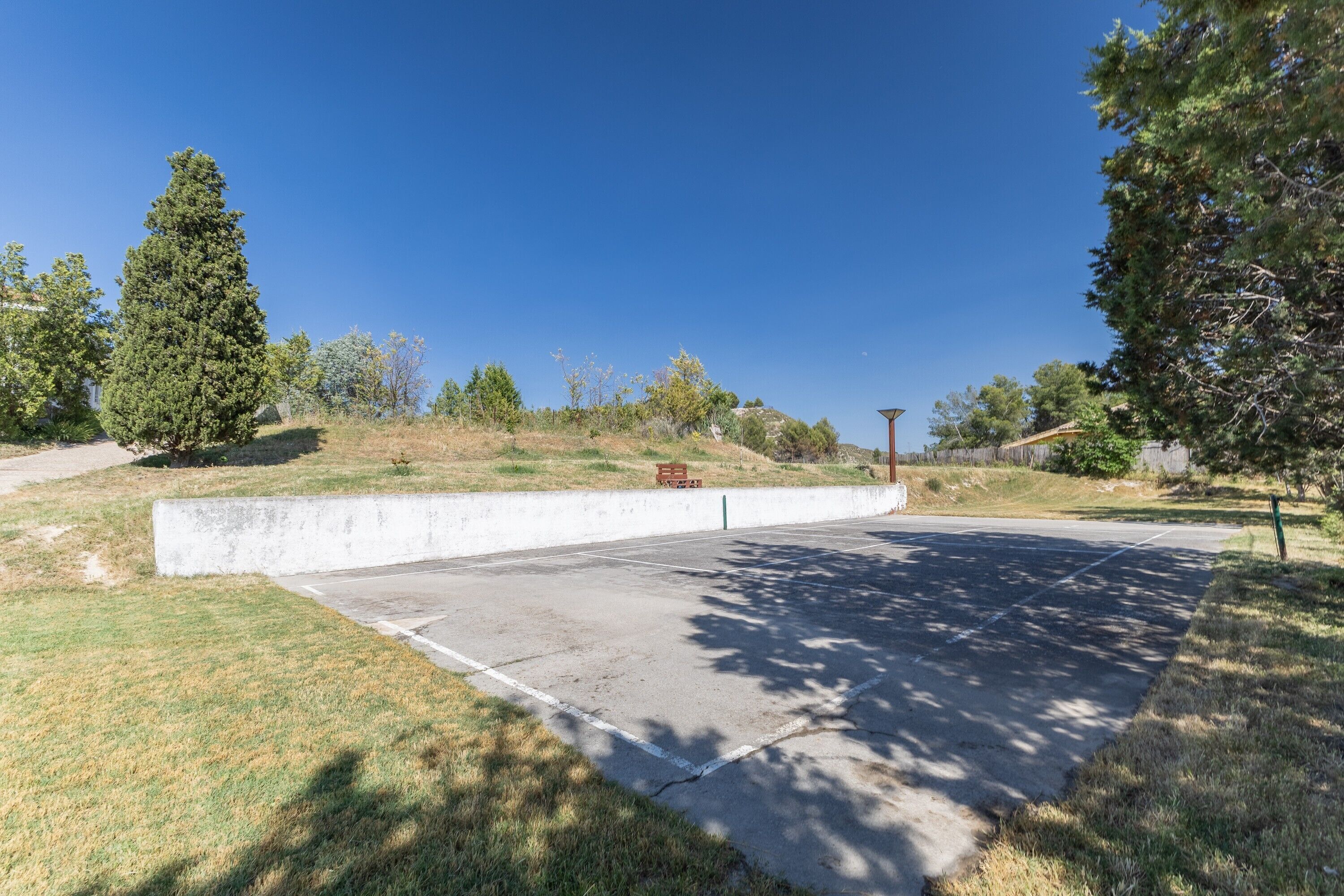 A concrete parking area with a bench, trees, and a clear blue sky.