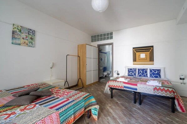 A room with two beds, a wooden floor, a bathroom with a shower, and a wall-mounted light fixture.