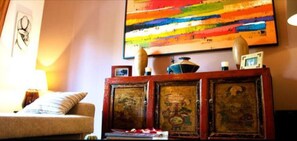 A room with a sofa, a cabinet with artwork, a colorful abstract painting on the wall, and a lamp.
