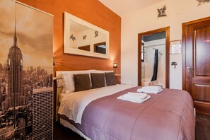 Une chambre d’hôtel avec un lit, un grand miroir, une affiche représentant un paysage urbain et une porte en bois.