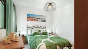 A bedroom with a bed, a nightstand, a table with a teapot and cups, a painting on the wall, and a hanging light fixture.