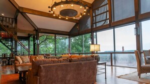 A spacious living area with a large sofa, a coffee table, and a chair. There is a staircase leading to an upper level, and large windows offering a view of the outdoors.