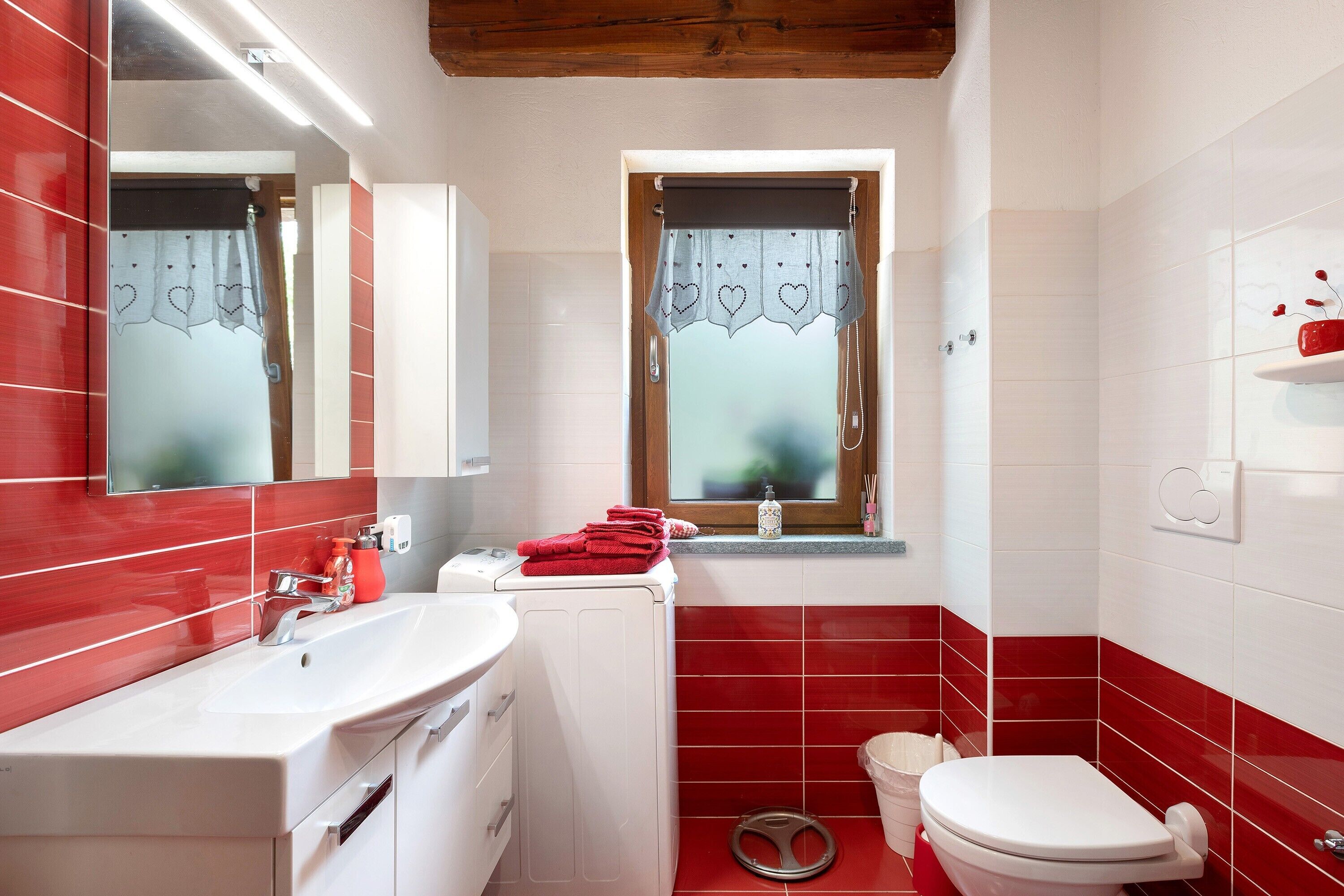 Une salle de bain avec un lavabo blanc, des murs recouverts de carreaux rouges, un miroir, une fenêtre avec un rideau et des toilettes.