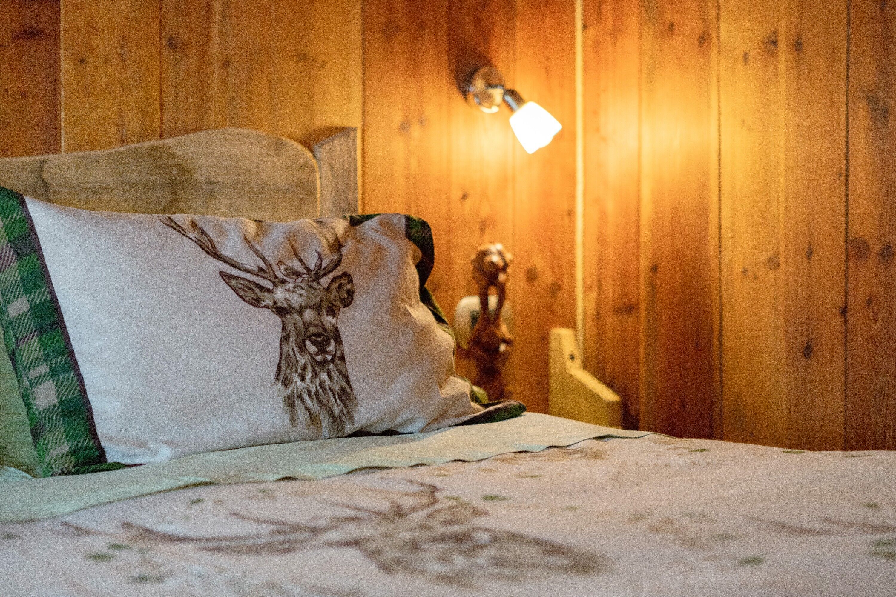 A bed with a deer pillow, a wooden headboard, a wall-mounted lamp, and a wooden nightstand with a figurine.