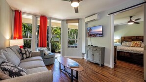 A modern living room with a sofa, coffee table, and a TV mounted on the wall. There is a sliding door leading to a balcony with red curtains.