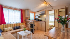 Una kitchenette compacta con microondas, estufa y pileta. Cuenta con una pequeña mesa de comedor con sillas, un sofá con cojines y un ventanal con cortinas.