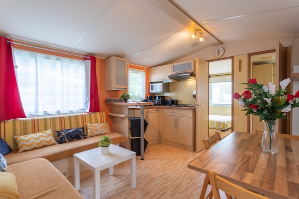 Une petite cuisine équipée comprenant un four à micro-ondes, une cuisinière et un évier. On y trouve également une petite table à manger avec des chaises, un canapé avec des coussins et une fenêtre avec des rideaux.