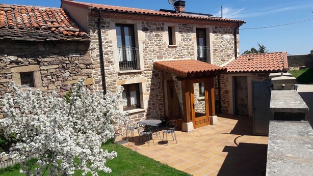 Un edificio de piedra con techo de tejas, un patio con mesa y sillas, y un árbol en flor.