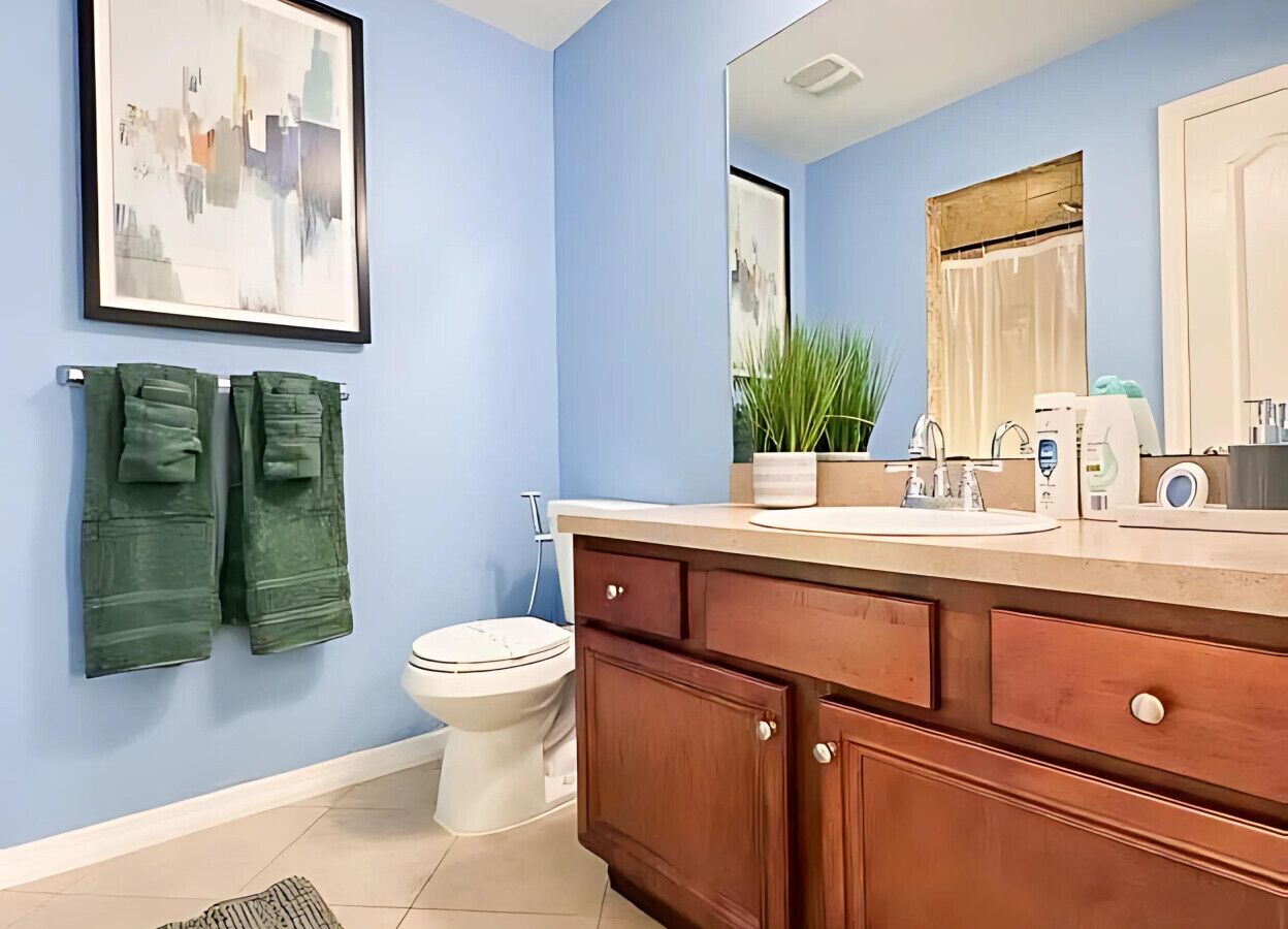 Une salle de bain avec un mur bleu, des toilettes blanches, un meuble-lavabo en bois avec un évier, et un tableau encadré au-dessus du miroir.